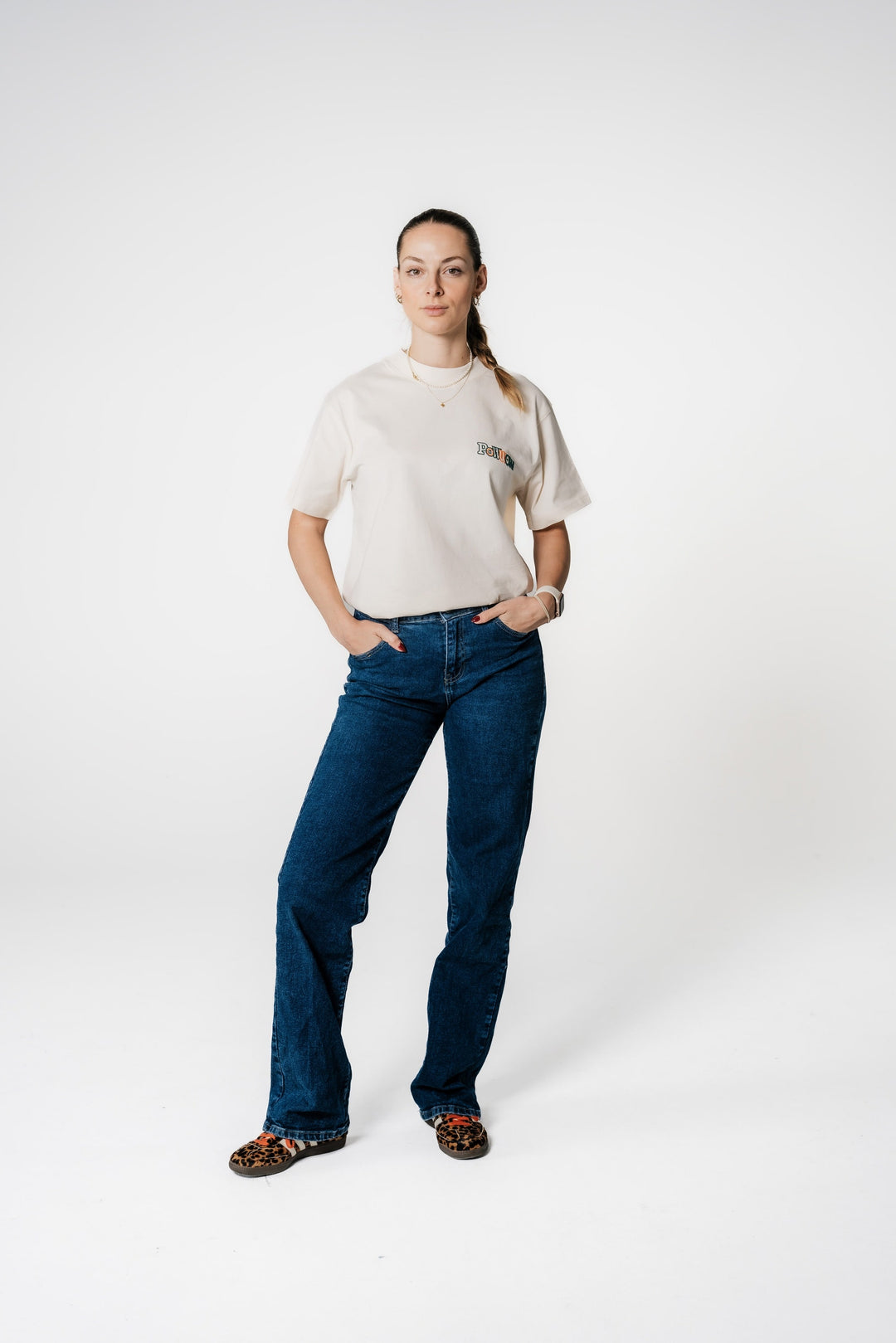 Model steht mit Händen in den Hosentaschen und trägt das beige POWWOW Classic Logo T-Shirt in CREAM (Off-White) mit kleinem Logo-Stick auf der Brust.