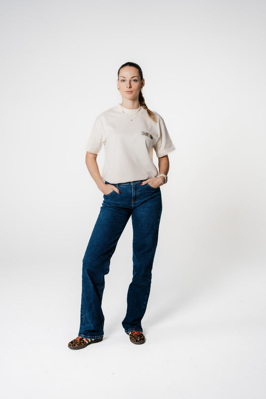 Model steht mit Händen in den Hosentaschen und trägt das beige POWWOW Classic Logo T-Shirt in CREAM (Off-White) mit kleinem Logo-Stick auf der Brust.