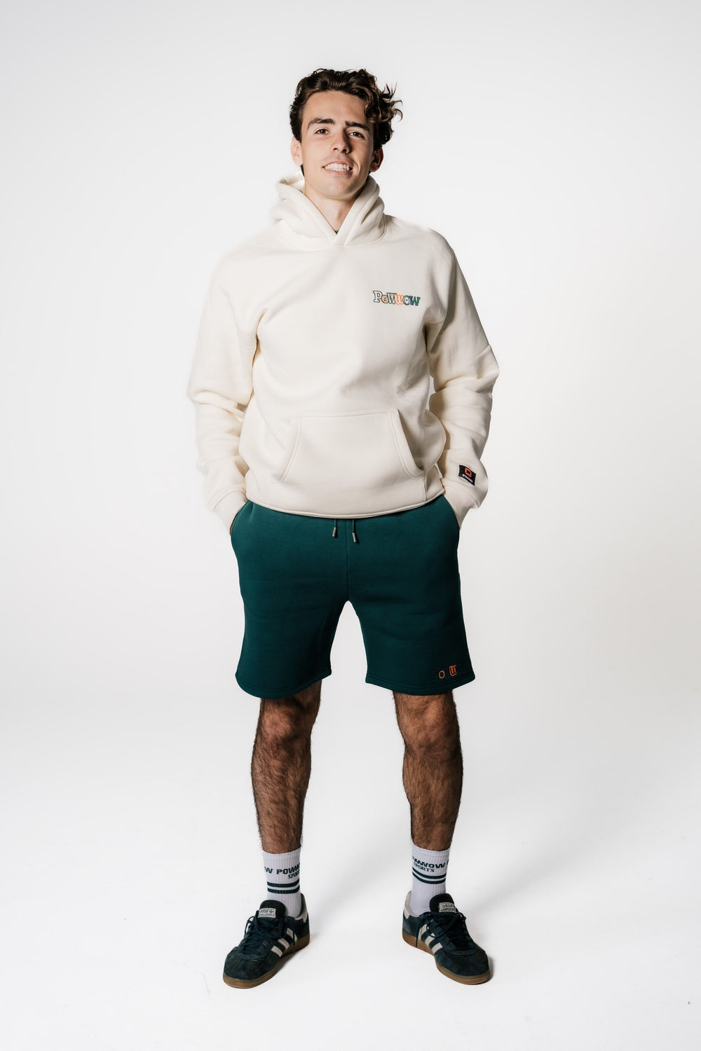Model trägt die dunkelgrünen POWWOW x Stanno Team Shorts zusammen mit dem cremefarbenen POWWOW Signature Hoodie.