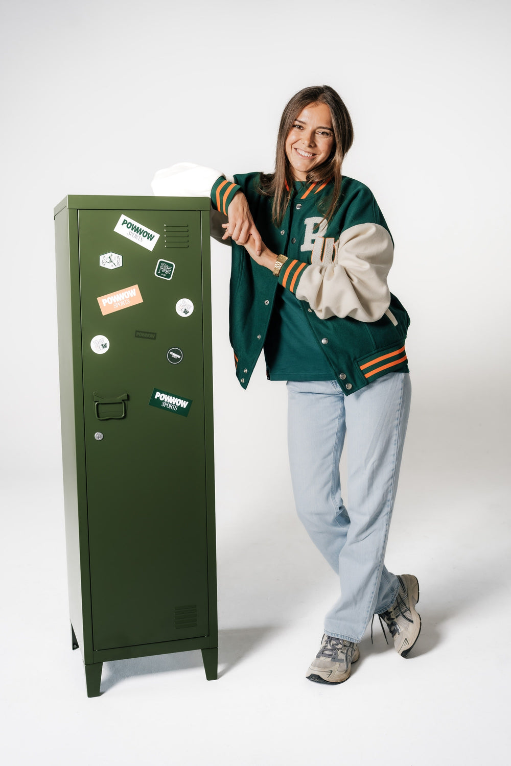 Model lehnt an einem grünen Spind und trägt die geschlossene POWWOW College-Jacke in Grün und Creme.