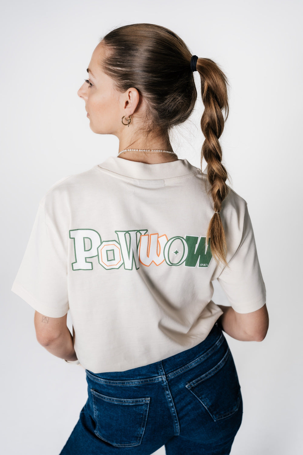Rückansicht eines Models, das beige POWWOW Backprint T-Shirt in CREAM trägt. Der große 'Powwow'-Schriftzug ist in Grün Green und Orange auf dem Rücken gedruckt.