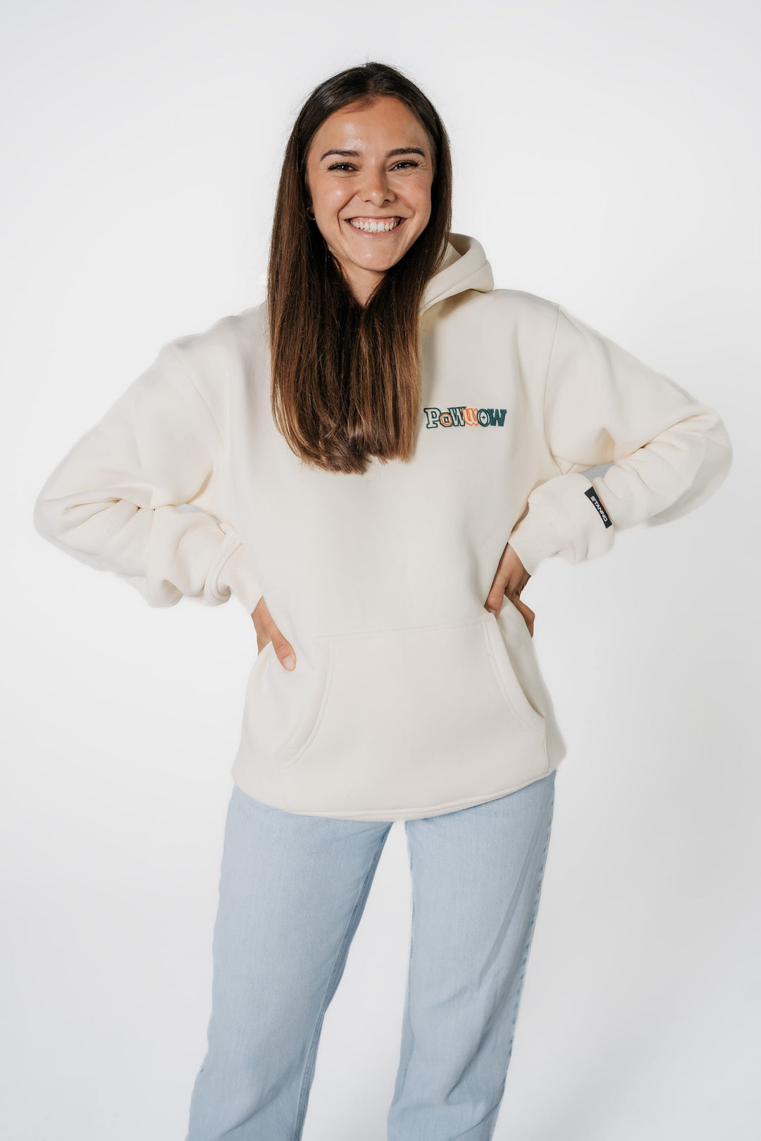 Model lächelt und trägt den beigen, cremefarbenen POWWOW Signature Hoodie CREAM mit kleinem Logo-Stick auf der Brust.