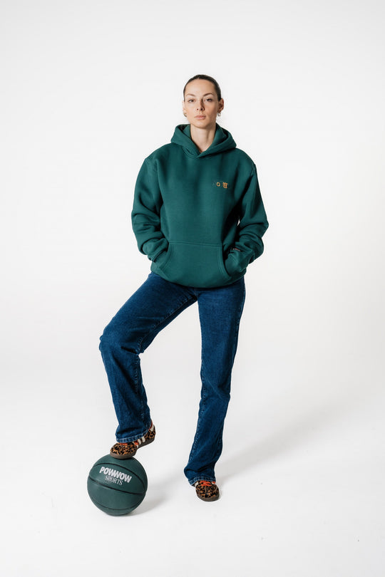 Model steht mit einem Fuß auf einem POWWOW Basketball und trägt den dunkelgrünen POWWOW Signature Hoodie GREEN.