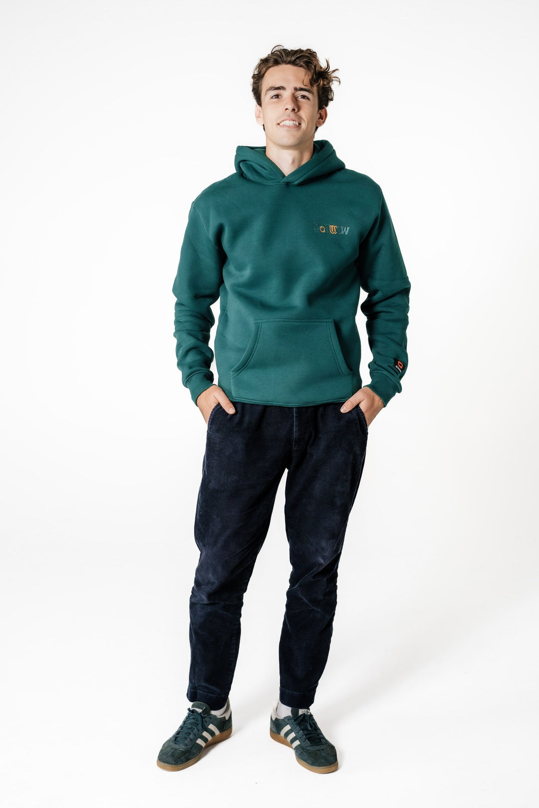 Model steht mit Händen in den Hosentaschen und trägt den dunkelgrünen POWWOW Signature Hoodie GREEN mit kleinem Logo-Stick auf der Brust.