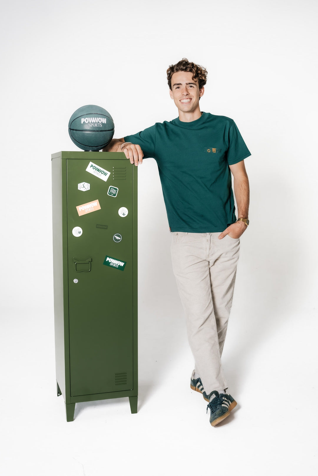 Model lehnt an einem grünen Spind und trägt das dunkelgrüne POWWOW Classic T-Shirt green mit kleinem Logo-Stick auf der Brust.