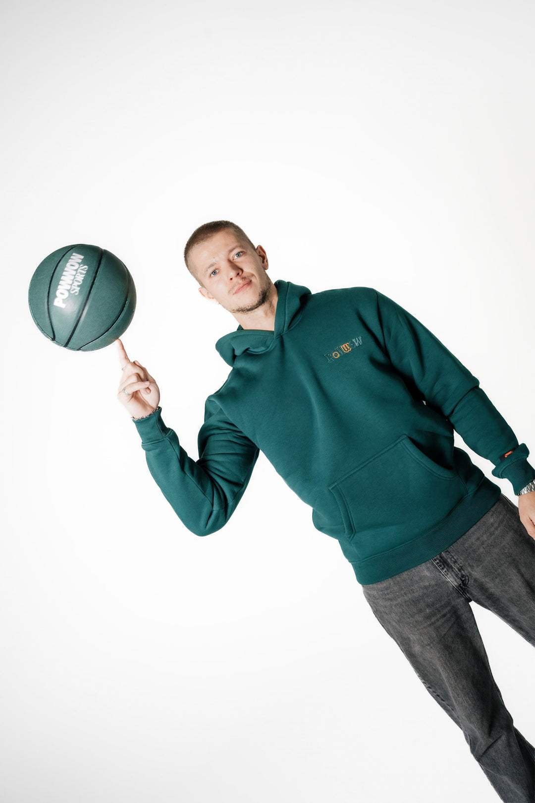 Model balanciert einen POWWOW Basketball auf dem Finger und trägt den dunkelgrünen POWWOW Signature Hoodie GREEN mit kleinem Brust-Logo.