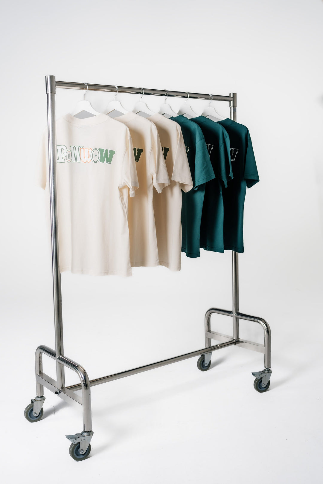 Die POWWOW Backprint Logo T-Shirts in CREAM und GREEN hängen auf einem Kleiderständer und zeigen den großen Logo-Print auf dem Rücken.